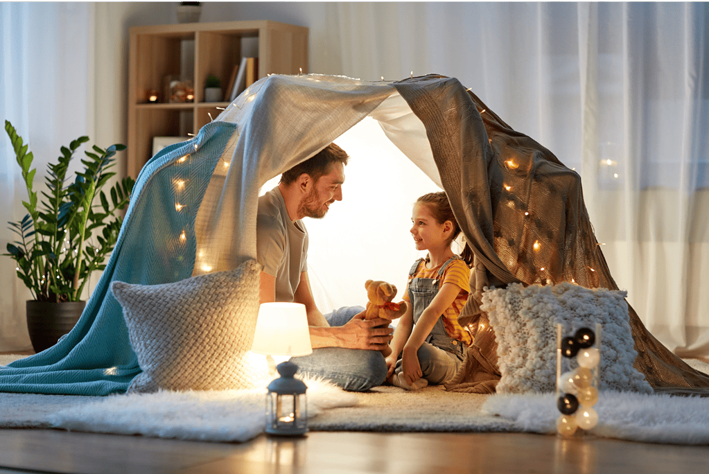 A father and daughter in a sheet fort with a teddy bear